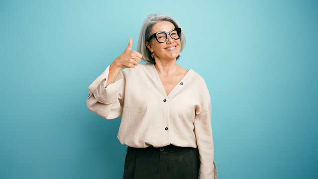Happy senior woman with gray hair and glasses smiling and showing thumbs up gesture, indicating approval or success