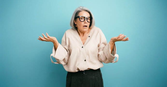 Elder woman shrugging shoulders with hands open, showing confusion and doubt over blue studio background