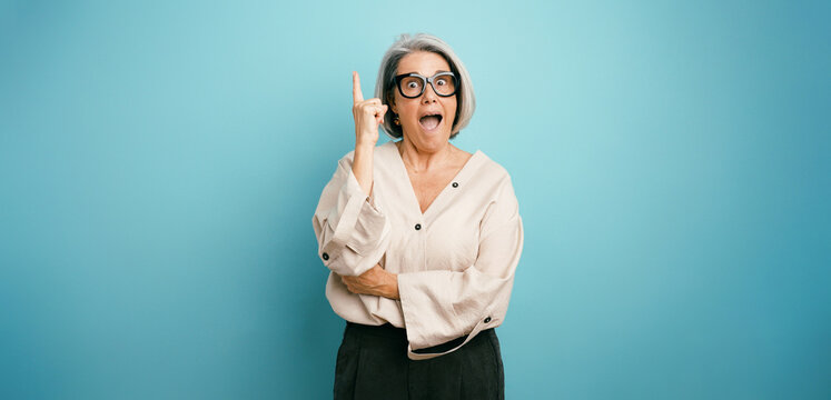 Mature woman with gray hair wearing glasses pointing finger up, expressing excitement and finding a new creative idea