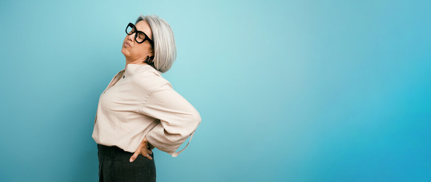 Senior woman suffering from lower back ache, touching her side with a pained expression standing against a blue background