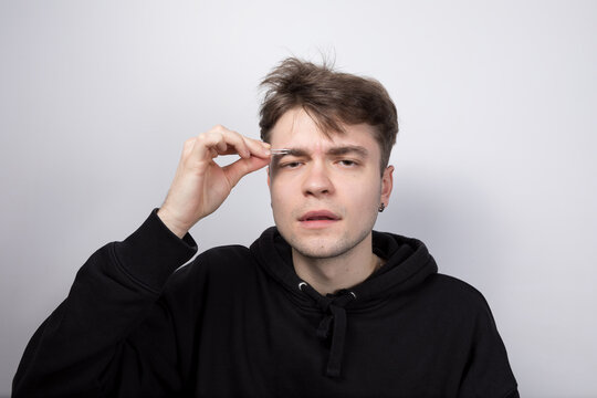 Thoughtful young man plucking his eyebrows with precision using tweezers in a casual black hoodie