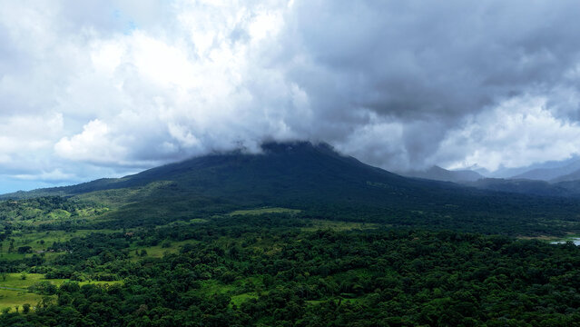 Arenal volcano peak covered by clouds, surrounded by dense tropical rainforest and green pastures in Costa Rica