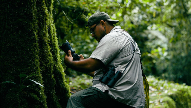 Man in outdoor clothing holding a camera with binoculars hanging, exploring the dense green mossy rainforest environment