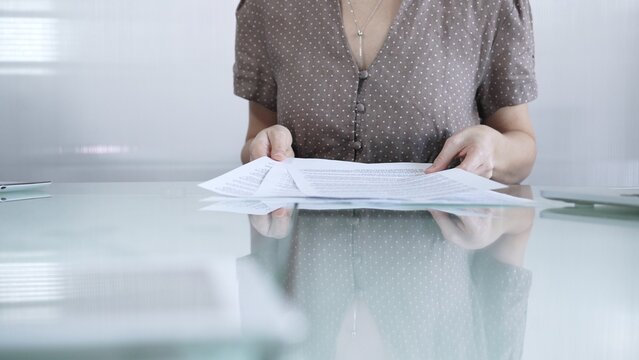 Businesswoman or accountant in brawn dotted blouse is going through paperwork while holding documents at a glass desk in a well lit, contemporary office setting. Business people concept