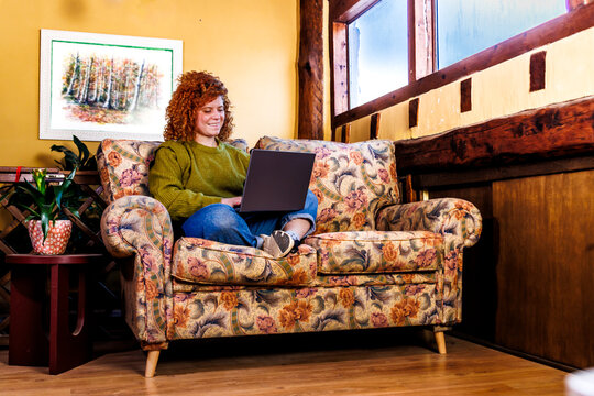 Woman with curly red hair working from home on laptop, sitting on armchair