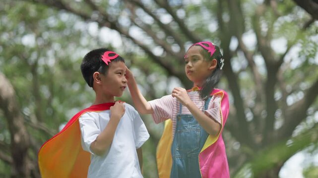 Cute Asian children playing superhero roleplay with colorful cape and mask running in outdoor green park. Kids imagination, creative future dream, and brave hero inspiration.