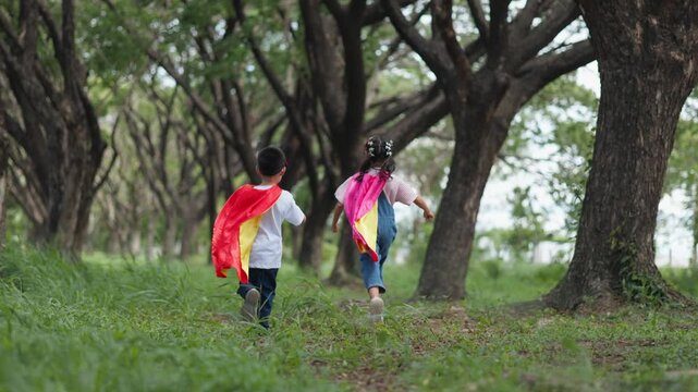 Cute Asian children playing superhero roleplay with colorful cape and mask running in outdoor green park. Kids imagination, creative future dream, and brave hero inspiration.