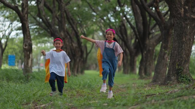 Active Asian children wearing superhero cape and mask having fun running in tree tunnel park. Kids imagination, creative future dream, and brave hero inspiration activity.