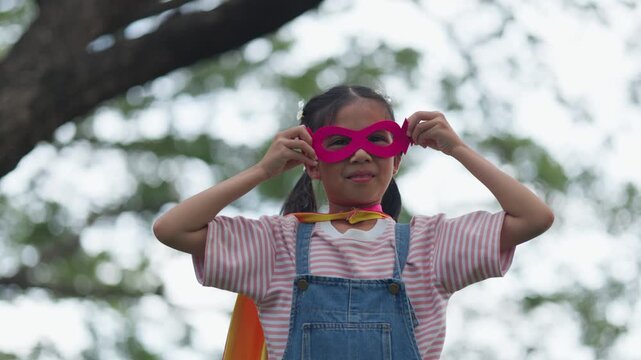 Cute Asian children playing superhero roleplay with colorful cape and mask running in outdoor green park. Kids imagination, creative future dream, and brave hero inspiration.