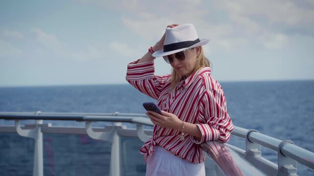 Girl enjoying cruise somewhere in ocean as she checks her smartphone on ship deck. Blonde woman in striped shirt and white fedora hat leans against railing with blue sea and cloudy sky behind her.
