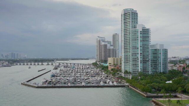 Port of Miami aerial footage showcasing marina filled with luxury yachts and boats. Modern high-rise condominiums line the waterfront with palm trees and green parks below cloudy skies.