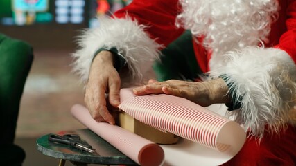 Fototapeta premium Santa Claus wraps presents in colorful paper after finishing the nice list and prepares the gifts for his Christmas journey to visit children.