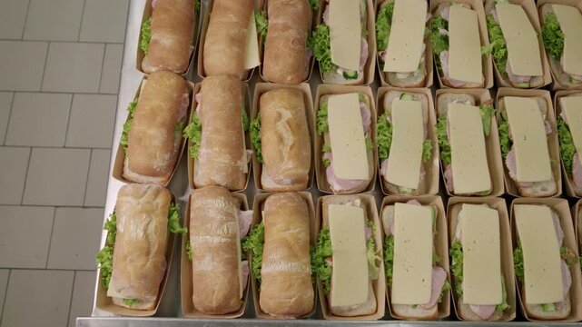 Hand in black glove places sandwich roll on top of prepared sandwich assembly line with lettuce and cheese in a commercial kitchen setting