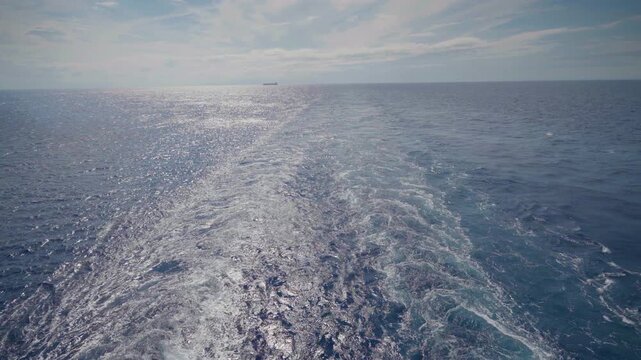 Foamy white ship wake creates a turbulent trail across deep blue ocean waters under partly cloudy sky. Distant cargo vessel silhouette visible on the horizon with sunlight reflecting on waves.