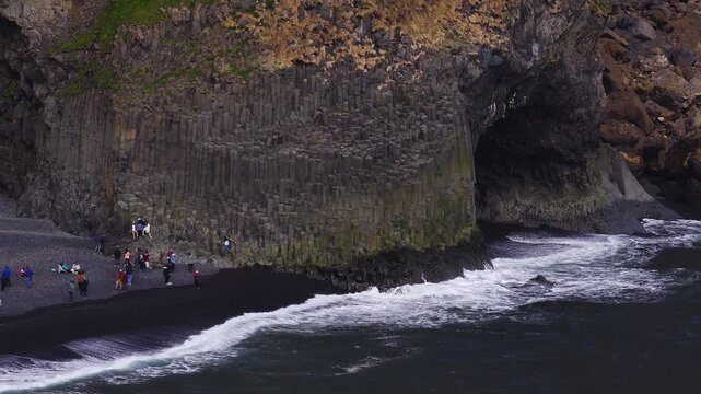 Drone flies over black pebble beach at Vik, Iceland with hexagonal basalt columns and dark cave, Atlantic waves break and foam, visitors walk, stand, and pose