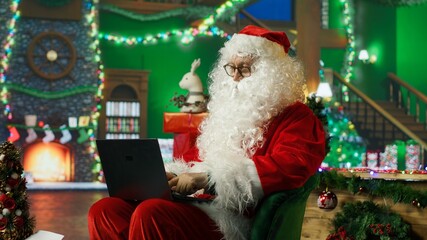 Fototapeta premium A smiling man dressed as Santa Claus reads a letter from children on a laptop and reviews their Christmas wish lists while wearing a cheerful red costume in a festive holiday setting.