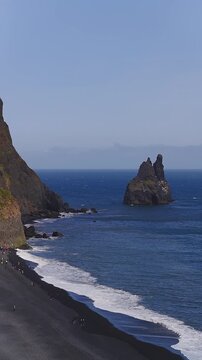 Aerial view of Reynisdrangar, basalt cliffs, and black sand near Vik, Iceland. Waves roll, visitors walk, daylight highlights columnar basalt in vertical frame.
