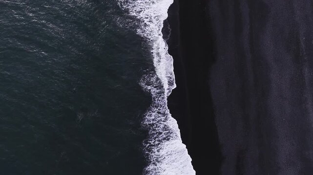 Aerial top down view shows waves breaking on Reynisfjara near Vik, Iceland. Rhythmic surf forms lace like foam on black sand under soft daylight, minimalist and abstract.