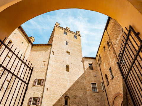 Palazzo dei Pio architecture standing tall in Carpi, Italy, showcasing the historic medieval tower and brick walls under a bright sky, framed by an ancient archway and metal fence