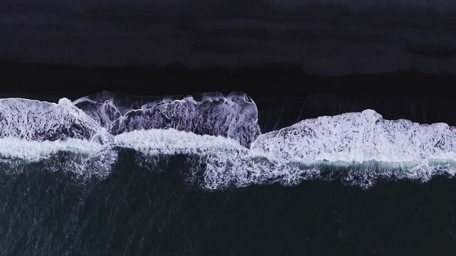 Aerial top down view of Vik black sand beach, Iceland. White surf rolls shoreward, foam patterns trace dark basaltic shore under muted overcast daylight.
