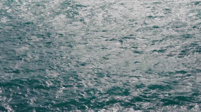 Choppy turquoise ocean water surface with small waves and bright sunlight reflections creating a shimmering pattern. Deep teal sea texture with natural movement and light.
