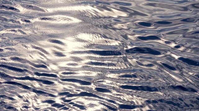 Sunlight creates brilliant silver reflections on calm water surface with gentle ripples and subtle wave patterns. Shimmering light dances across peaceful water creating mesmerizing abstract textures.