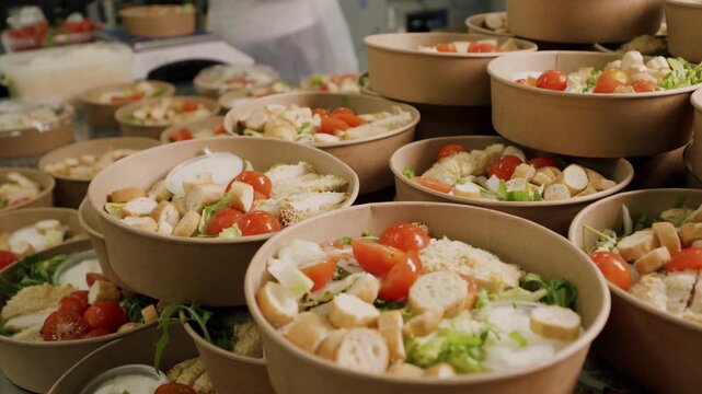 Multiple bowls of fresh salad with cherry tomatoes, croutons, and greens stacked on a table in a kitchen setting, ready for serving or distribution