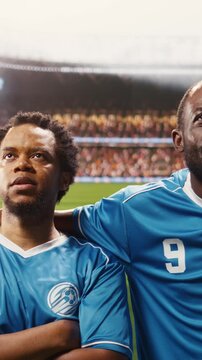 Vertical screen: Portrait of diverse football players in blue jersey listening to national anthem during pre-match ceremony, match day opening on pitch with team in lineup.