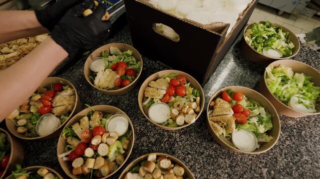 Preparation of fresh salads in brown bowls with cherry tomatoes, croutons, and dressing, as a gloved hand adds ingredients in a commercial kitchen setting