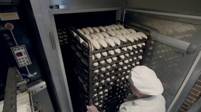 Baker in white uniform places trays of dough into a commercial oven, showcasing the process of preparing bread in a professional kitchen environment