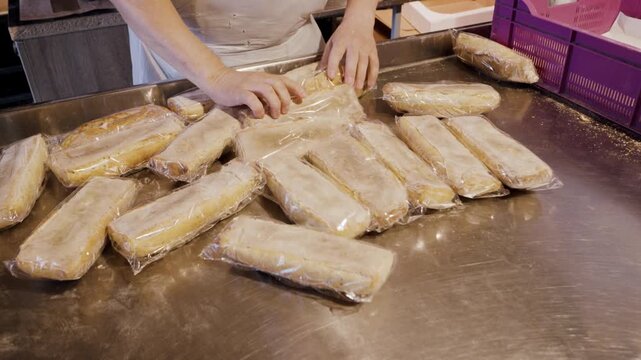 Bakery worker arranging packaged bread loaves on stainless steel table, with purple basket and kitchen equipment visible in the background