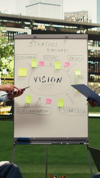 Vertical composition: Diverse business people explaining vision implementation with sticky notes, gesturing with open hands during outdoor presentation, informal discussion with coworkers.
