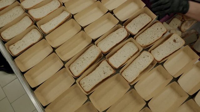 Worker in gloves places sliced bread into paper trays on a table, showcasing a systematic arrangement of loaves in a commercial kitchen environment