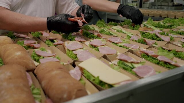 Caucasian female worker in gloves assembles sandwiches with lettuce, ham, and cheese in a commercial kitchen, surrounded by trays of prepared ingredients and bread rolls