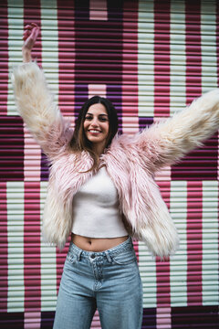 Cheerful young woman raising both arms in triumph against vibrant background, happy girl in pink faux fur coat celebrating success and happiness, authentic people lifestyle and sincere emotions.