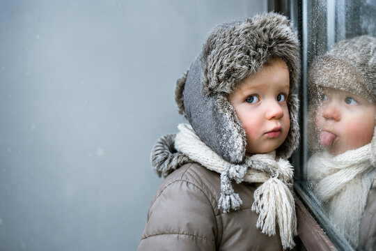 Thoughtful child in winter clothes looking through a fogged window at another playful child and ample space for text, cozy lifestyle concept in cold weather childlike innocence, banner for advertising