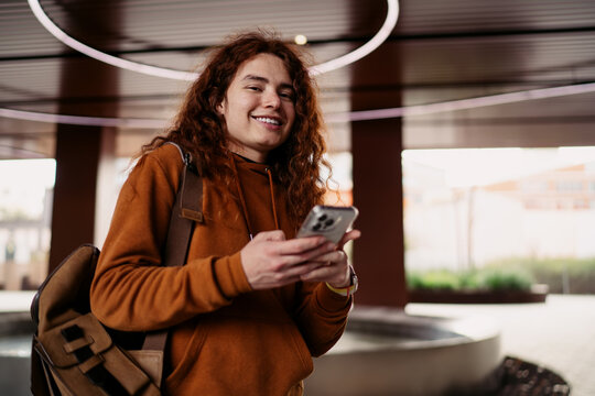 Happy young man with ginger curly hair smiling holding mobile phone, male student using smartphone application for social media, authentic people lifestyle, digital communication, Caucasian model.