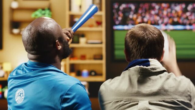 Tight framing shows football supporters eating popcorn and drinking beer while watching live match on TV as a group of friends hold fan props and enjoy snacks during a home watch party.