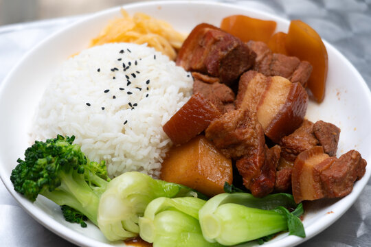 A delicious bowl of braised pork belly served with steamed white rice, bok choy, broccoli, pickled vegetables, and simmered radish. A comforting Asian meal featuring tender meat and vibrant greens.