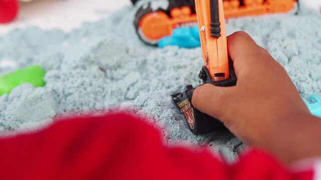 Close-up of a child's hand playing with an orange toy excavator, digging in light blue kinetic sand, emphasizing childhood creative play and sensory development.