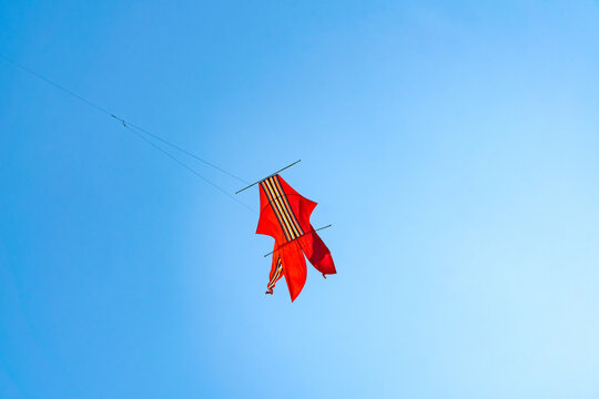 A traditional Balinese bebean or f kite, vibrant red with a central striped pattern, soars high against a clear blue sky. The bamboo structure and tail are visible. Captured during a festival in Bali.