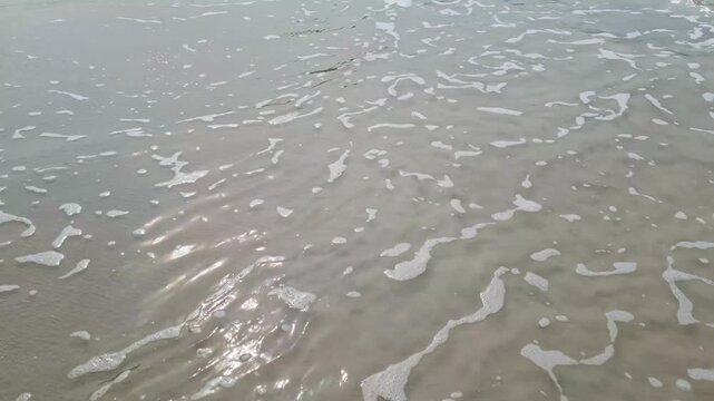 Detailed close-up shot of rich white sea foam and delicate bubbles spreading over wet sand as ocean waves retreat.