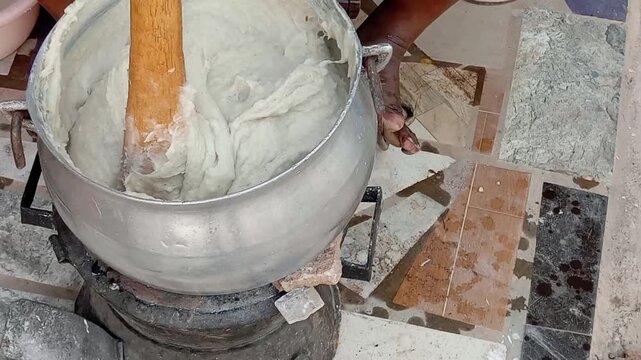 African preparing banku in large metal pot on coal pot at home, house. Stirring with wooden spoon. Ghanaian traditional food made by mixing liquid mixture of corn and cassava dough. Food preparation.