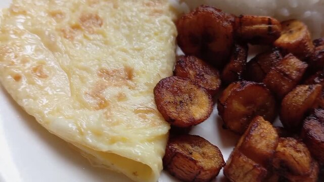 Braised rice with fried small pieces of plantain, egg and red grinded pepper sauce served on white plate. African cuisine, restaurant menu and food serving. Food served on black and white tablecloth.