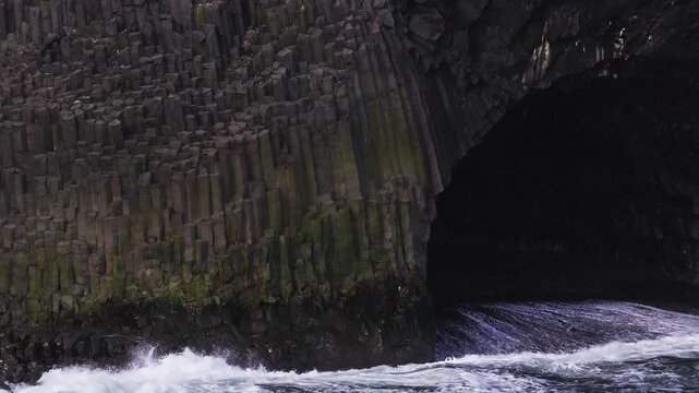 Aerial view shows hexagonal basalt columns and a dark sea cave near Vik, Iceland, as Atlantic waves crash, white foam churns, and moss streaks mark the jagged waterline.