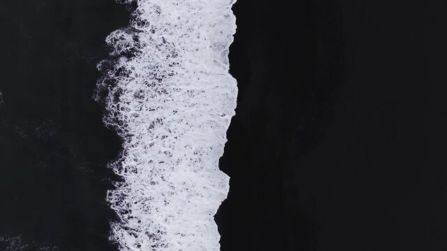 Aerial top down view of surf meeting black sand at Reynisfjara near Vik, Iceland. Slow waves surge and retreat, foam patterns form under soft daylight.