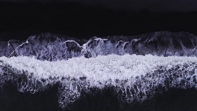 Aerial view shows waves pound Reynisfjara black sand near Vik, Iceland. Swells roll, break, and recede, leaving lacy foam bands in a high contrast monochrome style.
