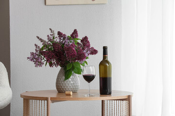 Obraz premium Red wine bottle and glass with a vase of purple lilac flowers on a wooden table indoors, Set indoors with a white wall and a sheer white curtain in the background, illuminated by natural light