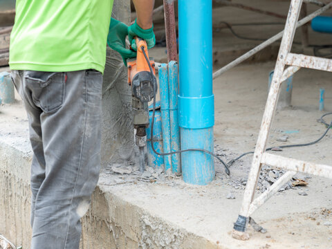 a construction worker using an electric jackhammer to break concrete ground next to blue PVC plumbing pipes.