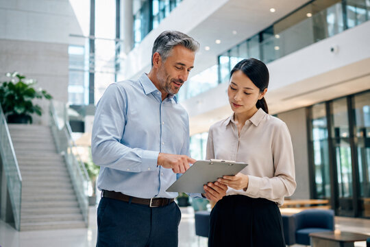 Senior male mentor and young female employee reviewing a clipboard in a spacious corporate atrium, depicting workplace training, guidance, onboarding, and knowledge sharing.
